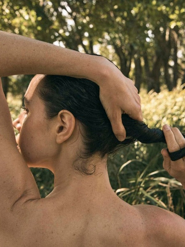 Rear view of a person gathering dark, wet hair at the nape of the neck with both hands, outdoors among green plants.