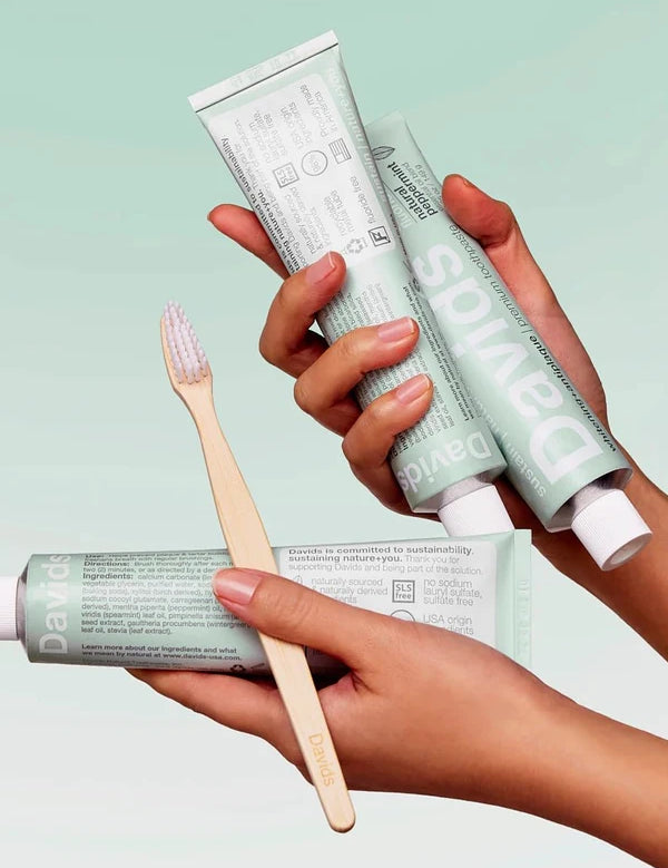 Hands holding Davids Peppermint Toothpaste tubes with a bamboo toothbrush