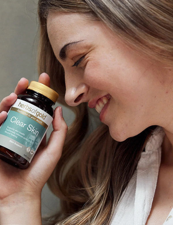 Smiling woman holding the Herbs of Gold Clear Skin bottle beside her cheek