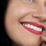 A smiling woman with smooth skin and bright white teeth wears vibrant red Hurraw! Rouge à Lèvres Plantcolor No 1 (4.8g) lipstick. “Hurraw! Rouge à Lèvres Plantcolor No 1” appears in the lower left, highlighting this plant-based lip colour.