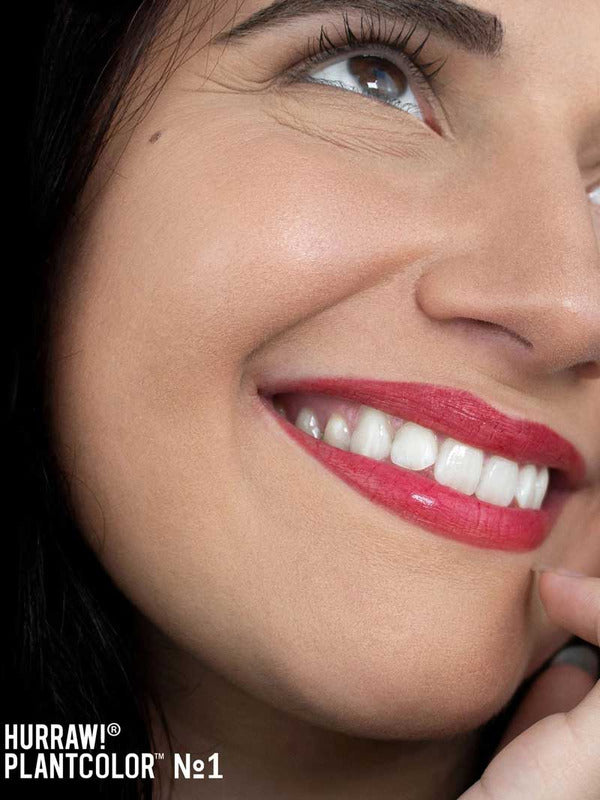 A smiling woman with smooth skin and bright white teeth wears vibrant red Hurraw! Rouge à Lèvres Plantcolor No 1 (4.8g) lipstick. “Hurraw! Rouge à Lèvres Plantcolor No 1” appears in the lower left, highlighting this plant-based lip colour.