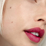 Close-up of a fair-skinned person with blonde hair, blue eye, and a cheek beauty mark wearing vibrant pinkish-red Hurraw! Rouge à Lèvres Plantcolor No 3 4.8g vegan lipstick. Text: “Hurraw! Plantcolor №3” in the bottom left corner.