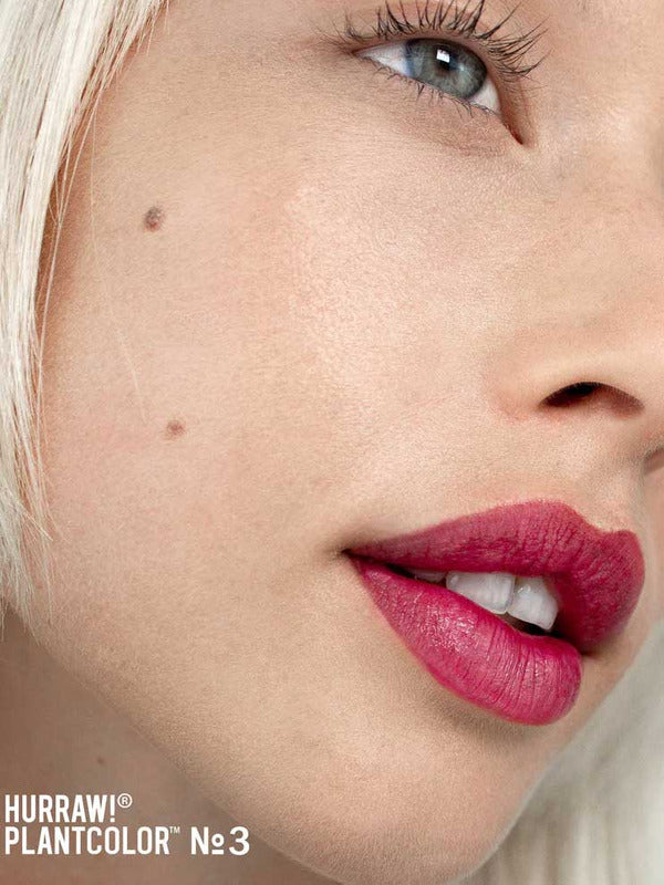 Close-up of a fair-skinned person with blonde hair, blue eye, and a cheek beauty mark wearing vibrant pinkish-red Hurraw! Rouge à Lèvres Plantcolor No 3 4.8g vegan lipstick. Text: “Hurraw! Plantcolor №3” in the bottom left corner.