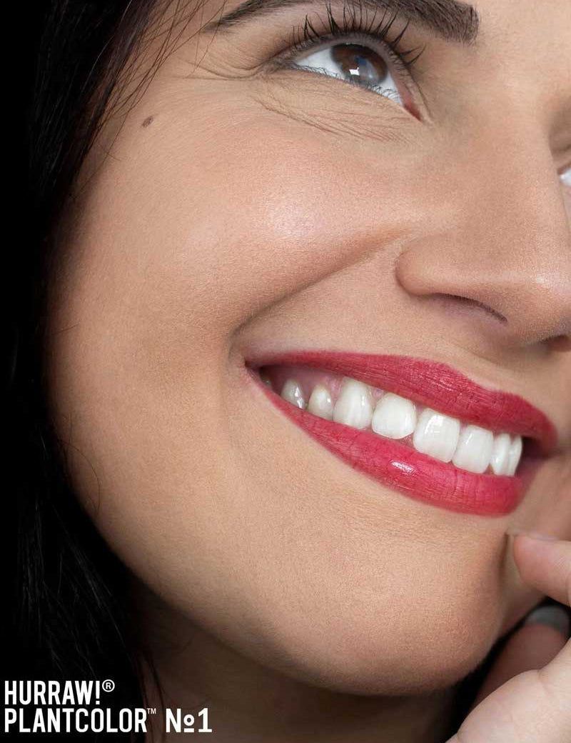 A smiling woman with smooth skin and bright white teeth wears vibrant red Hurraw! Rouge à Lèvres Plantcolor No 1 (4.8g) lipstick. “Hurraw! Rouge à Lèvres Plantcolor No 1” appears in the lower left, highlighting this plant-based lip colour.