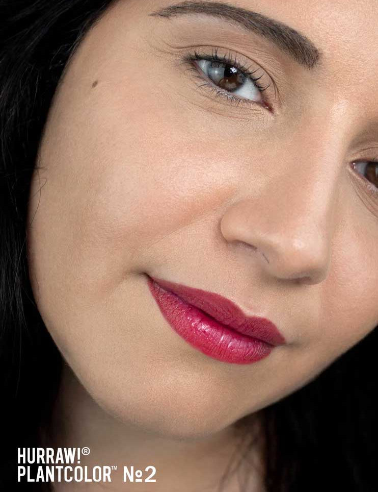 Close-up of a person with fair skin, dark hair, and dark eyebrows wearing bright pink Hurraw! Rouge à Lèvres Plantcolour No 2 4.8g lipstick. Text in the bottom left reads HURRAW! PLANTCOLOR™ №2.