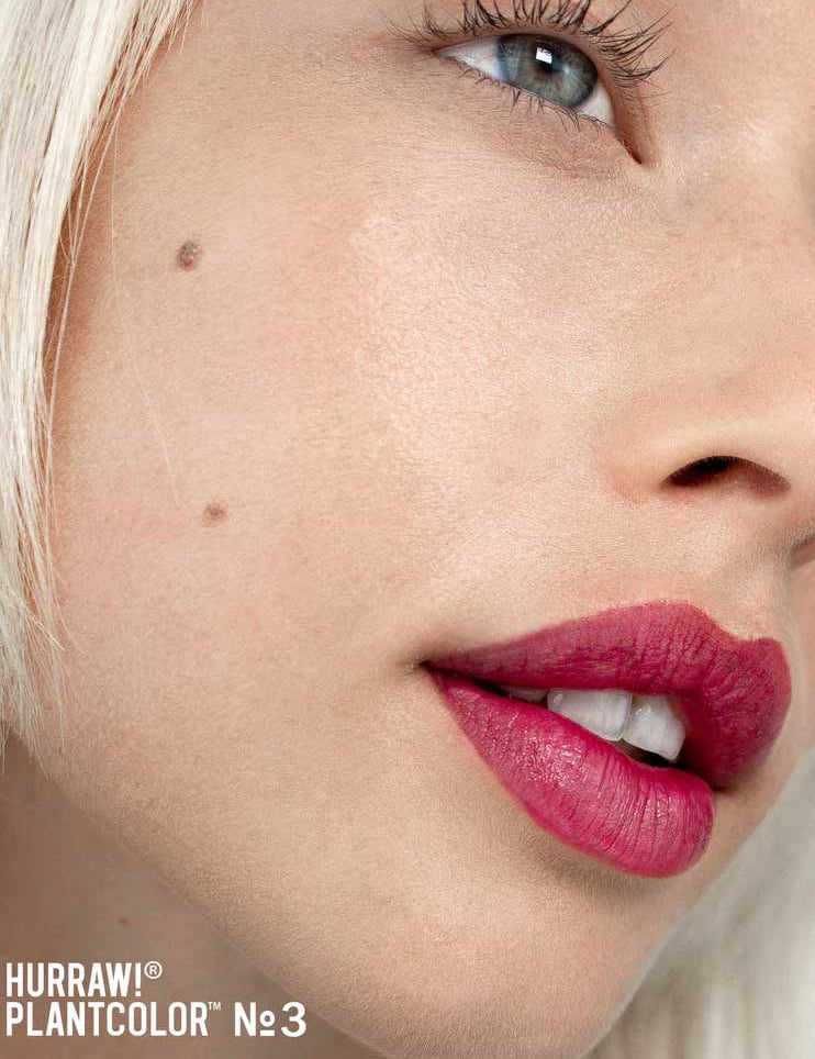 Close-up of a fair-skinned person with blonde hair, blue eye, and a cheek beauty mark wearing vibrant pinkish-red Hurraw! Rouge à Lèvres Plantcolor No 3 4.8g vegan lipstick. Text: “Hurraw! Plantcolor №3” in the bottom left corner.