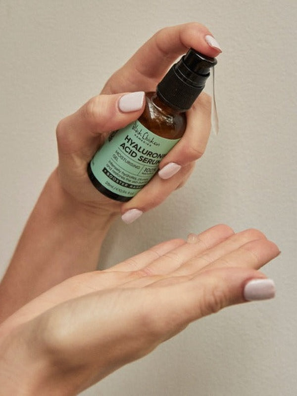 One hand holds an amber Hyaluronic Acid Serum bottle with a mint-green label, with a finger on the black pump above the other open palm.