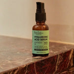 An amber Hyaluronic Acid Serum pump bottle with a mint-green label stands on a dark reddish-brown marble ledge against a beige wall.