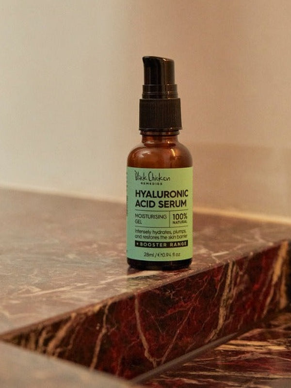 An amber Hyaluronic Acid Serum pump bottle with a mint-green label stands on a dark reddish-brown marble ledge against a beige wall.