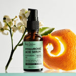 An amber Hyaluronic Acid Serum bottle with a black pump and mint-green label stands beside white flowers, a green leaf and a curled strip of orange peel.