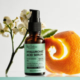 An amber Hyaluronic Acid Serum bottle with a black pump and mint-green label stands beside white flowers, a green leaf and a curled strip of orange peel.