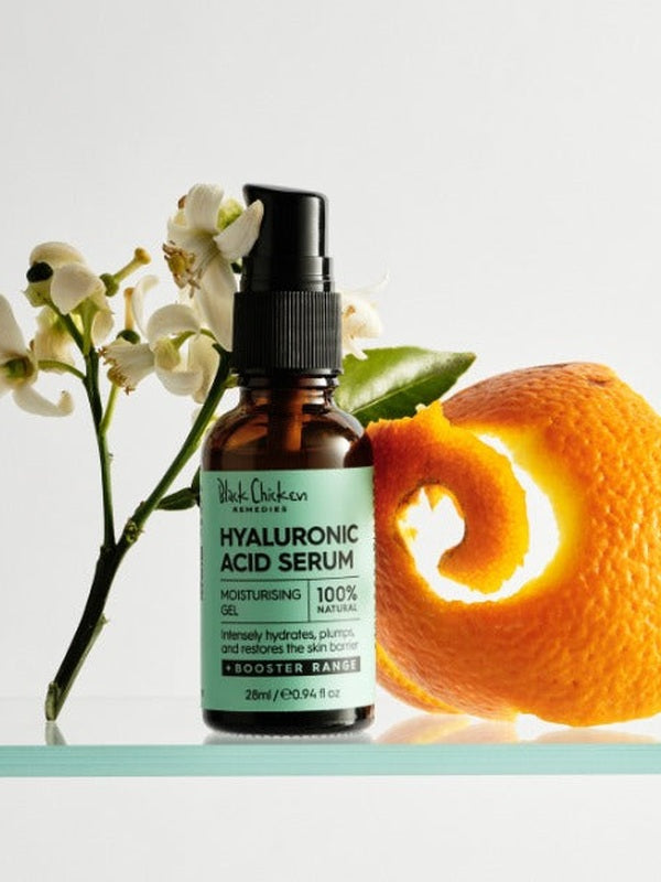 An amber Hyaluronic Acid Serum bottle with a black pump and mint-green label stands beside white flowers, a green leaf and a curled strip of orange peel.