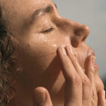 A woman closes her eyes and massages her cheeks with her fingertips in warm sunlight.