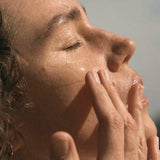 A woman closes her eyes and massages her cheeks with her fingertips in warm sunlight.