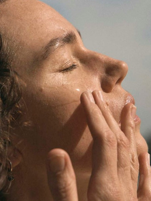 A woman closes her eyes and massages her cheeks with her fingertips in warm sunlight.