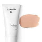 A white tube of Dr Hauschka Foundation 30ml is placed next to a beige natural foundation swatch, both offering medium coverage, on a white background.