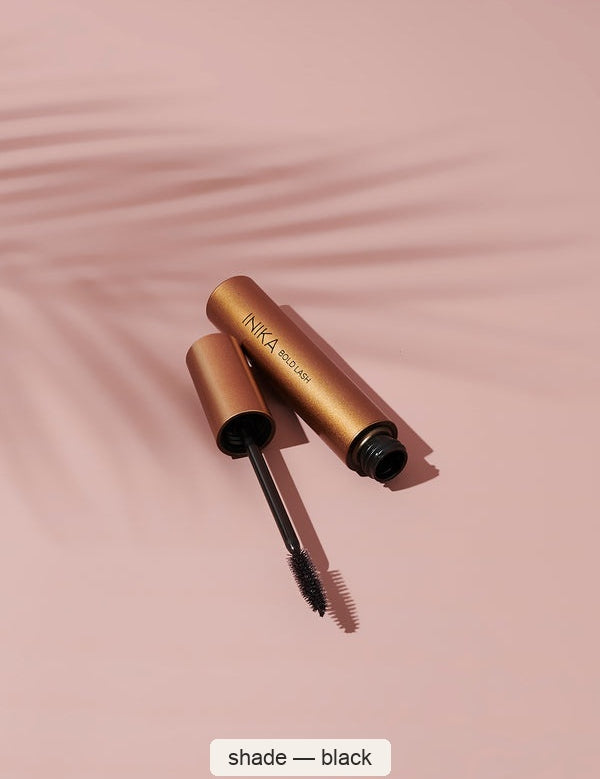 Open bronze INIKA Bold Lash mascara tube and a black-coated bristle wand resting diagonally on a pink surface with a palm-leaf shadow. The caption identifies the shade as Black.