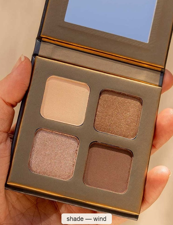 Hand holding an open gold INIKA Wind eyeshadow quad with four cream, bronze, taupe and brown pans beneath a rectangular mirror. The caption identifies the shade as Wind.