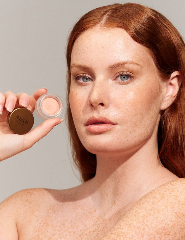 Freckled red-haired model holding an open pot of peach INIKA Organic Lip and Cheek Cream in Morning beside her face.