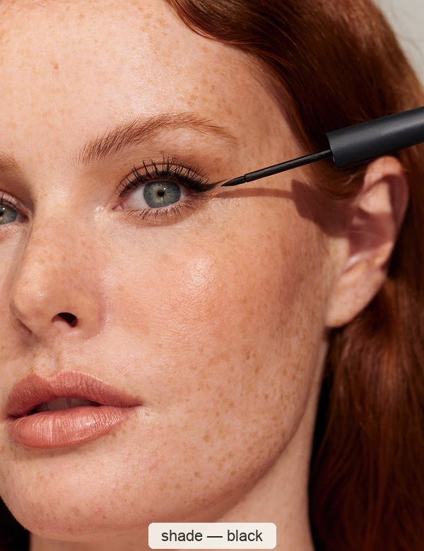 Close-up of a freckled person with red hair applying fine black eyeliner along the outer upper lash line with a pointed brush. The caption identifies the INIKA eyeliner shade as Black.