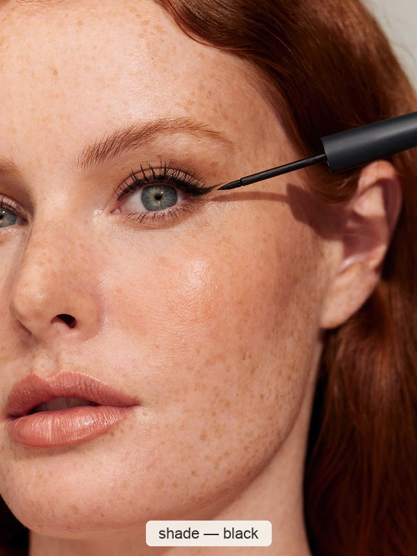 Close-up of a freckled person with red hair applying fine black eyeliner along the outer upper lash line with a pointed brush. The caption identifies the INIKA eyeliner shade as Black.