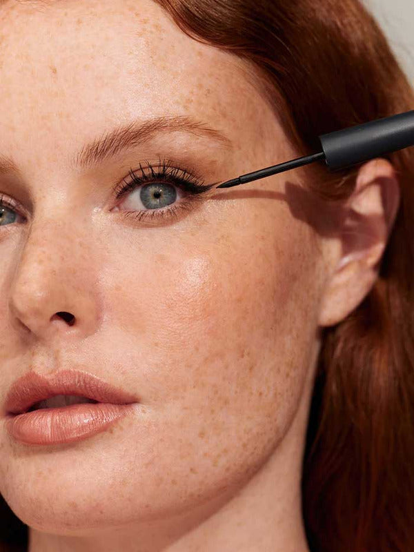 Close-up of a freckled model applying black INIKA Organic Liquid Eyeliner precisely along the upper lash line.