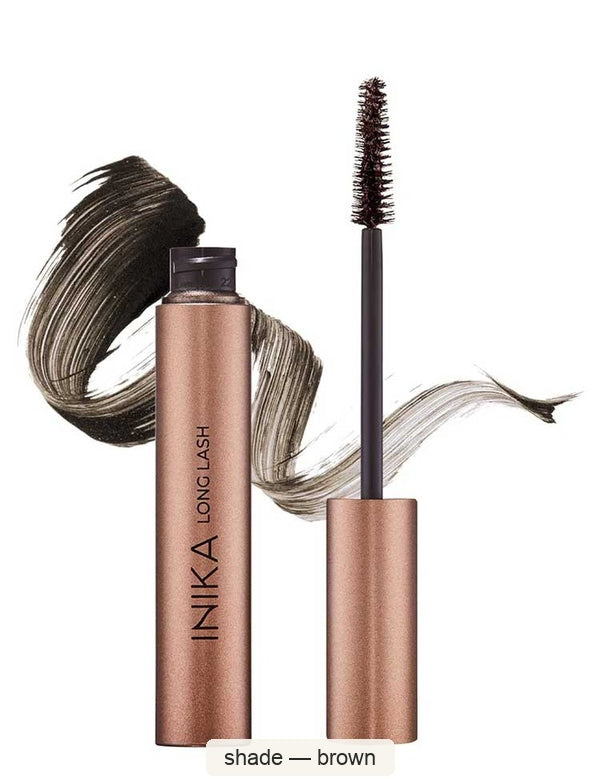 Open bronze INIKA Long Lash mascara tube beside an upright dark-bristled wand, with a sweeping brown mascara swatch behind them. The caption identifies the shade as Brown.