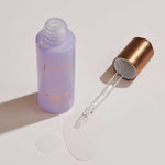 Open lavender INIKA Organic Bakuchiol Serum bottle and gold dropper arranged beside a pool of clear serum on a pale surface.