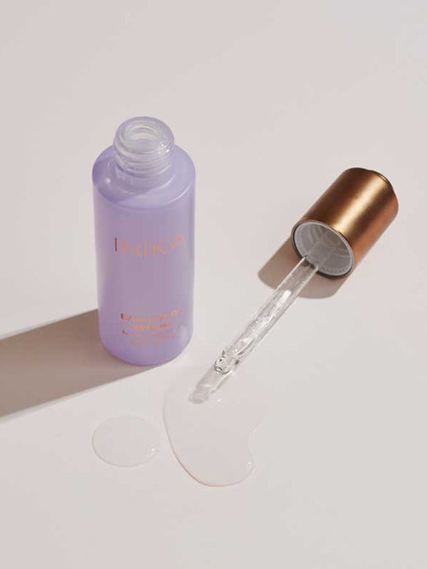 Open lavender INIKA Organic Bakuchiol Serum bottle and gold dropper arranged beside a pool of clear serum on a pale surface.