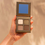 Hand holding an open INIKA Organic Eyeshadow Quad in Wind, showing four coordinated neutral matte and shimmer shades against a warm beige background.