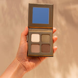 Hand holding an open INIKA Organic Eyeshadow Quad in Wind, showing four coordinated neutral matte and shimmer shades against a warm beige background.
