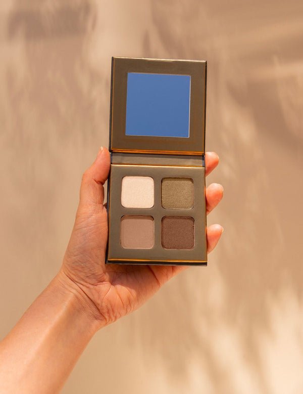 Hand holding an open INIKA Organic Eyeshadow Quad in Wind, showing four coordinated neutral matte and shimmer shades against a warm beige background.