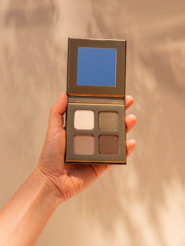 Hand holding an open INIKA Organic Eyeshadow Quad in Wind, showing four coordinated neutral matte and shimmer shades against a warm beige background.