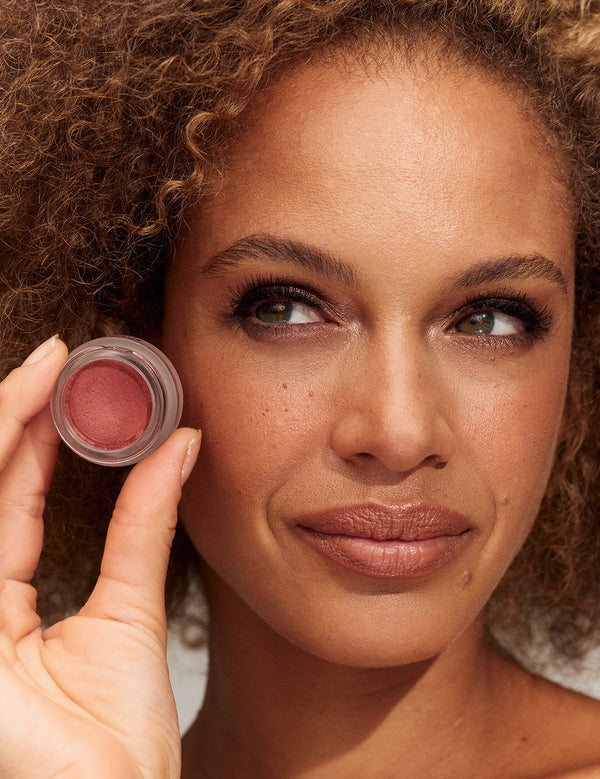 Person with curly light brown hair and rose-toned lips holding an open pot of deep rosy INIKA lip and cheek cream beside their face.