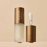 A frosted bottle of INIKA Organic Lip Oil - Collagen + Squalane Clear 5ml with a bronze cap is displayed; the cap stands beside the bottle, exposing a sponge-tip applicator. The backdrop features a soft beige tone.