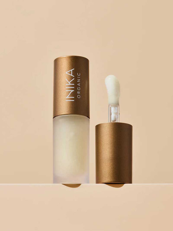 A frosted bottle of INIKA Organic Lip Oil - Collagen + Squalane Clear 5ml with a bronze cap is displayed; the cap stands beside the bottle, exposing a sponge-tip applicator. The backdrop features a soft beige tone.