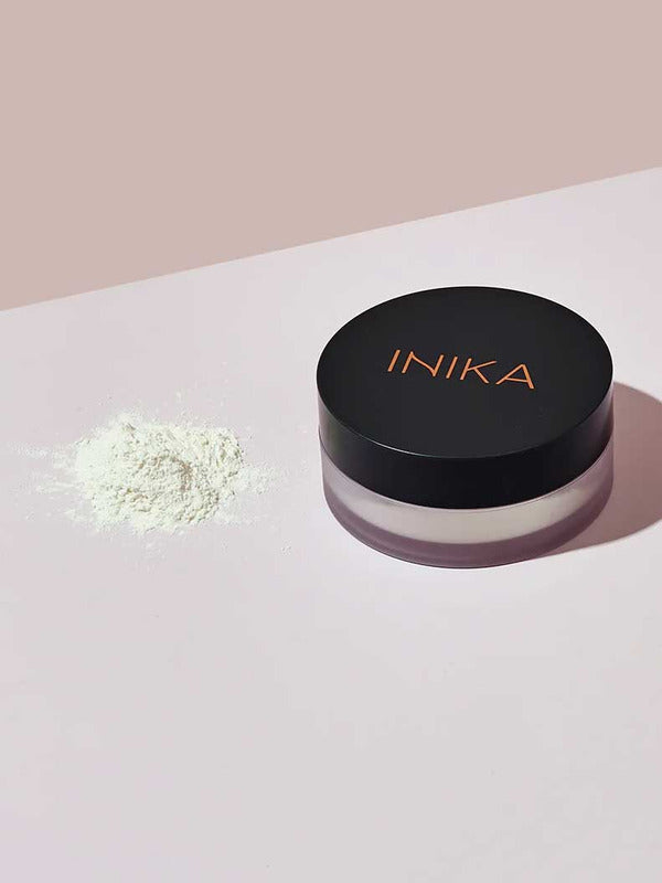 A small pile of loose white INIKA Organic Mattify Mineral Setting Powder 7g sits next to its round container with a black lid labeled "INIKA" in copper, placed on a light pink and white surface.