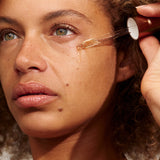 Close-up of a person with light brown curls holding a glass dropper beside their cheek, with a golden bead of INIKA Phyto-Active Face Oil at its tip.