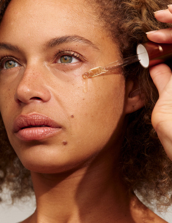 Close-up of a person with light brown curls holding a glass dropper beside their cheek, with a golden bead of INIKA Phyto-Active Face Oil at its tip.