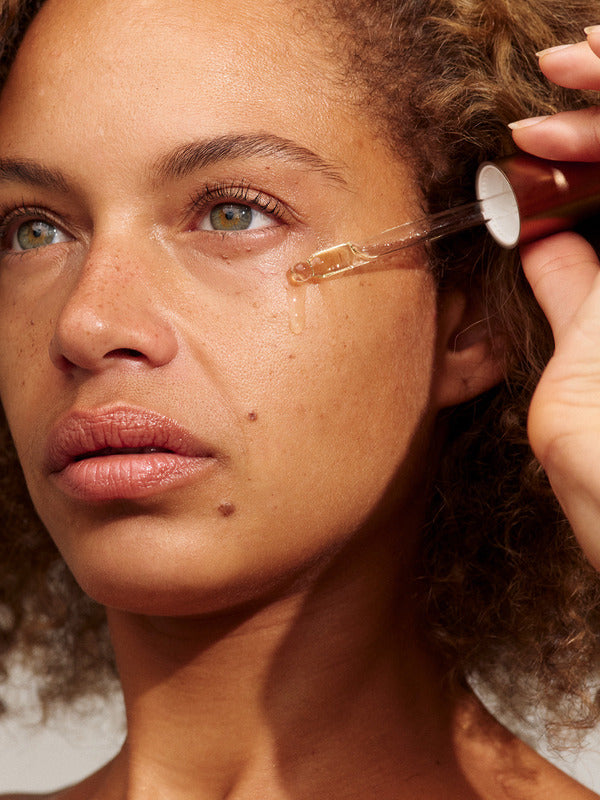 Close-up of a person with light brown curls holding a glass dropper beside their cheek, with a golden bead of INIKA Phyto-Active Face Oil at its tip.