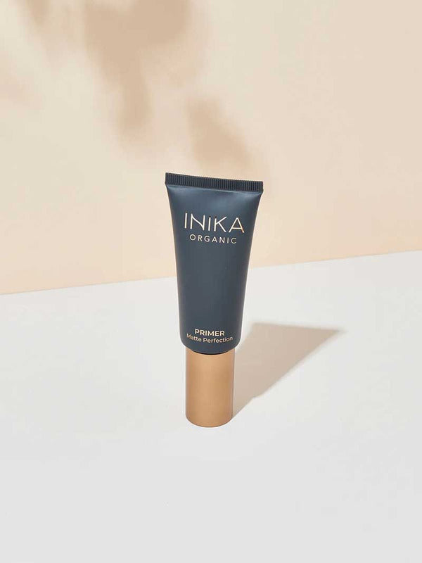 A 30ml tube of INIKA Organic Primer Matte Perfection, featuring a dark matte body and gold cap, stands upright on a white surface against a beige background.