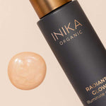 A bottle of INIKA Organic Radiant Glow Veil Primer 30ml rests beside a swatch of shimmery, light beige liquid on a neutral background.