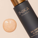 A bottle of INIKA Organic Radiant Glow Veil Primer 30ml rests beside a swatch of shimmery, light beige liquid on a neutral background.