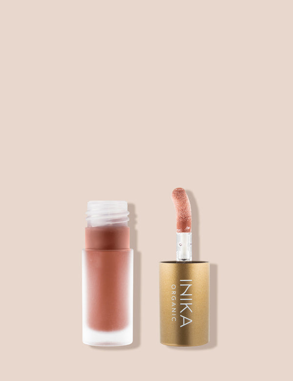 Open frosted INIKA tinted lip oil bottle containing peach-pink oil beside its upright sponge-tip applicator in a gold cap, on a beige background.