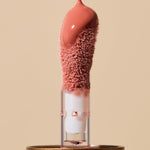 Glossy coral-pink INIKA lip oil pouring onto a fuzzy sponge applicator above a gold cap, forming a thick droplet over the tip. The caption identifies the shade as Blossom.
