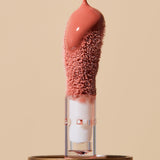 Glossy coral-pink INIKA lip oil pouring onto a fuzzy sponge applicator above a gold cap, forming a thick droplet over the tip. The caption identifies the shade as Blossom.