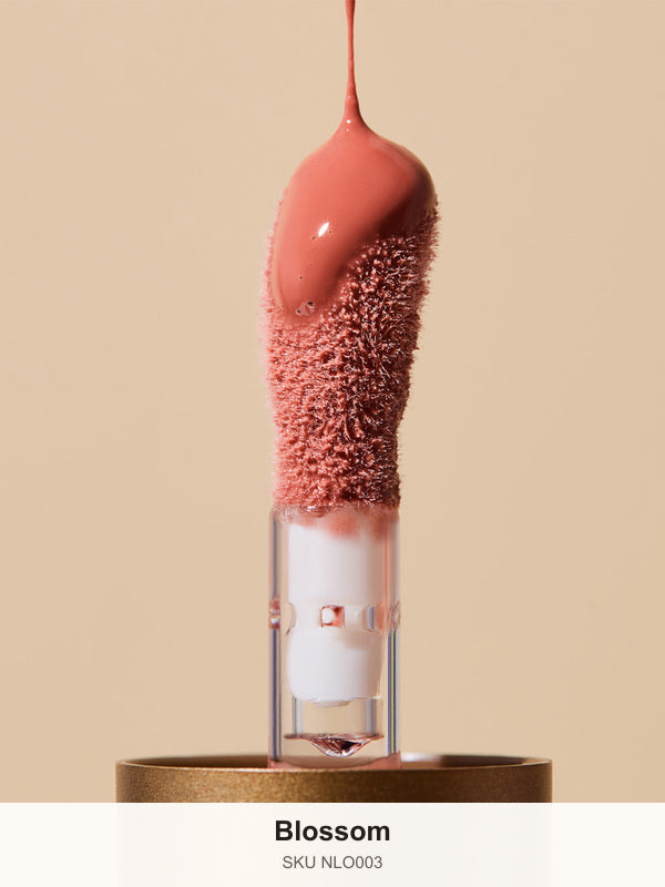 Glossy coral-pink INIKA lip oil pouring onto a fuzzy sponge applicator above a gold cap, forming a thick droplet over the tip. The caption identifies the shade as Blossom.