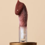 Glossy reddish-brown INIKA lip oil pouring onto a fuzzy sponge applicator above a gold cap, forming a thick droplet over the tip. The caption identifies the shade as Cinnamon.