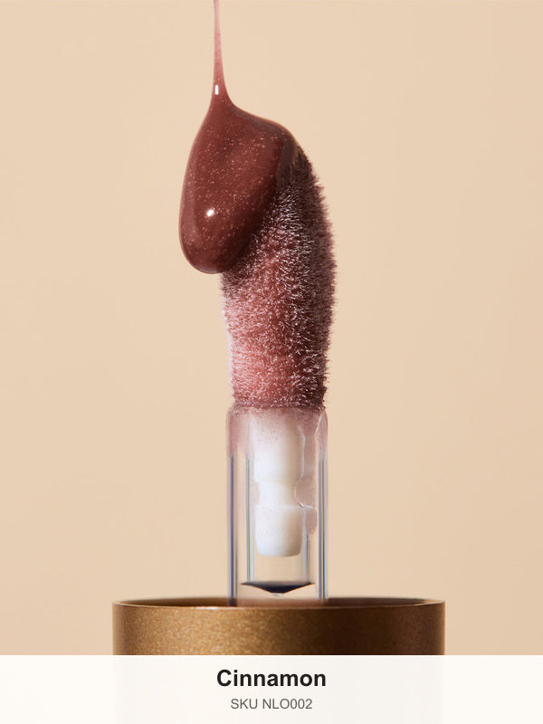 Glossy reddish-brown INIKA lip oil pouring onto a fuzzy sponge applicator above a gold cap, forming a thick droplet over the tip. The caption identifies the shade as Cinnamon.