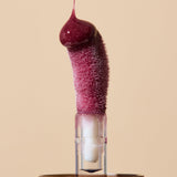Glossy deep berry INIKA lip oil pouring onto a fuzzy sponge applicator above a gold cap, forming a rounded droplet over the tip. The caption identifies the shade as Mulberry.